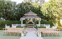 A garden wedding ceremony venue featuring a gazebo, string lights, hedges and draped linen with candles and floral arrangements lining the aisle